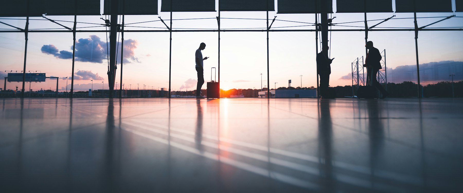 Travelers with luggage silhouetted against the windows of an airport terminal at sunrise